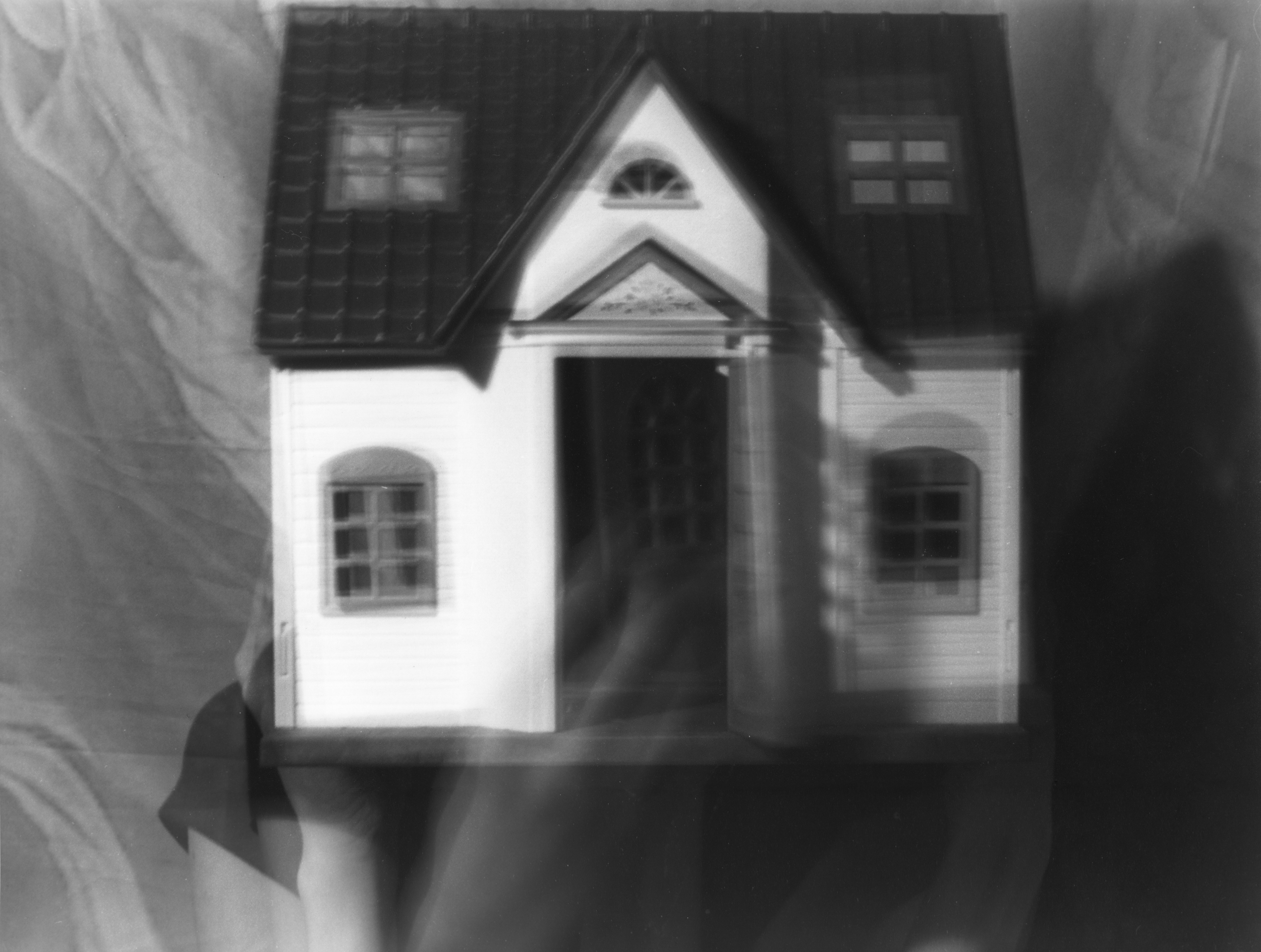 Black and white gelatin silver print of a woman whose face is completely covered by a dollhouse. Two images overlay one another, one in which the woman holds the dollshouse still over her head; the other in which the front door is being opened by her hand